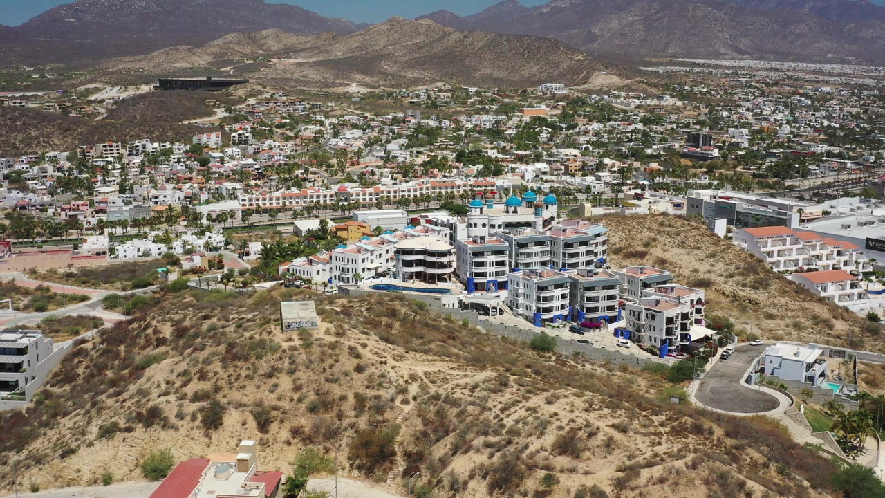 Aerial view in San Jose del Cabo with dry brush in the foreground, apartment blocks near the lower center, city streets and homes across the valley, mountains behind, and no visible people.