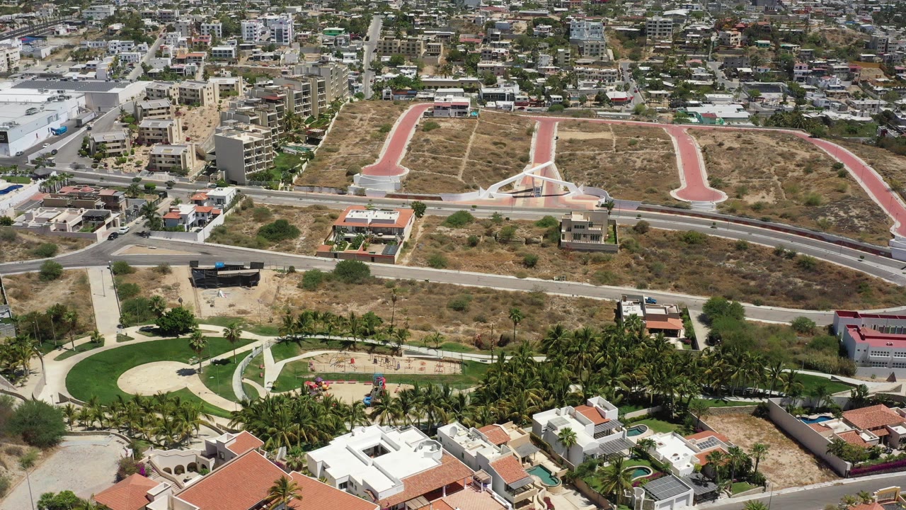 Aerial frame from San Jose del Cabo showing terracotta-roofed homes in the foreground, palm greenery, curving roads, dry open land, red paved paths, and no visible people.