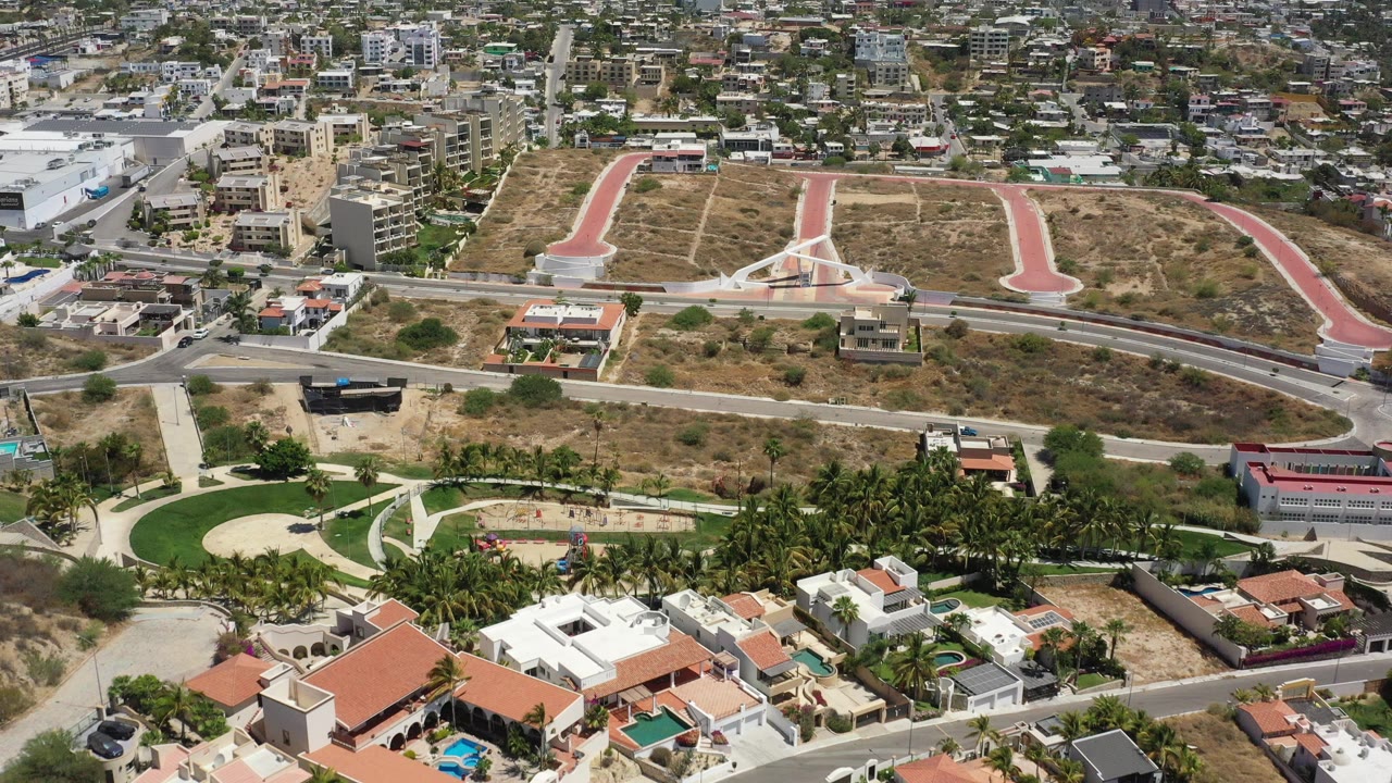 Opening aerial frame over San Jose del Cabo showing dry undeveloped lots above walled homes, pools, palm landscaping, and a distant city background with no visible people.