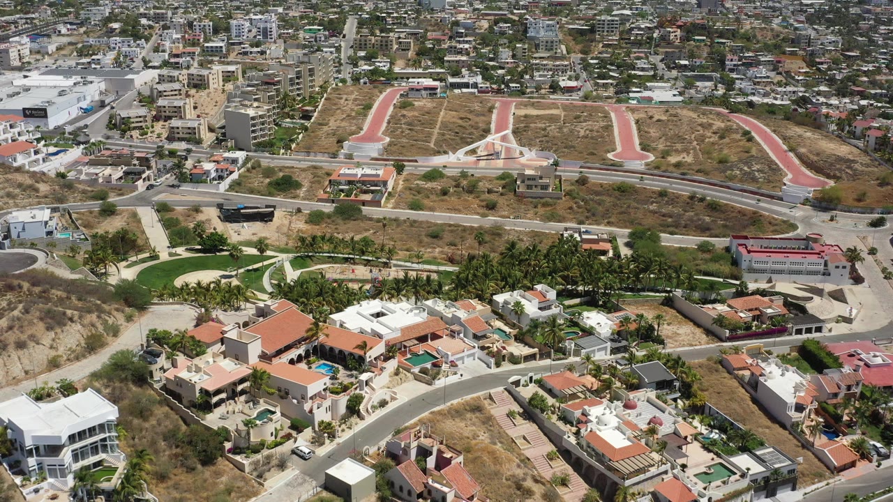 Aerial frame from San Jose del Cabo with villas and palms in the foreground, a dry slope and roadway through the middle, and compact city blocks across the background, with no visible people.