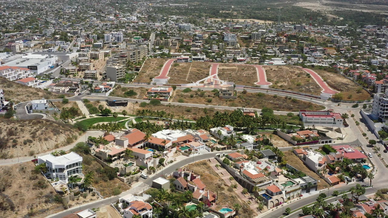 Opening aerial frame over San Jose Del Cabo with foreground hillside homes, red paved paths across a dry slope, distant town blocks, mountains, and no visible people.