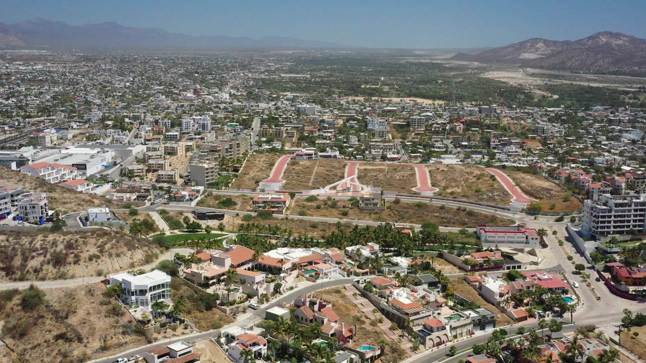 Wide aerial view of a sunlit desert city with hills, roads, homes, and mountains.