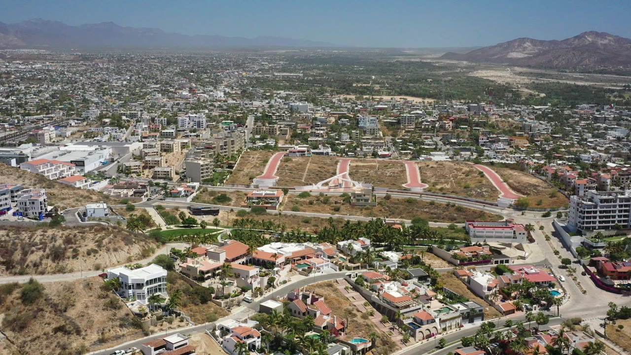 A high aerial frame of San Jose del Cabo, Mexico, showing residential slopes in front, a dense town grid beyond, desert ground, mountains on the horizon, and no visible people.