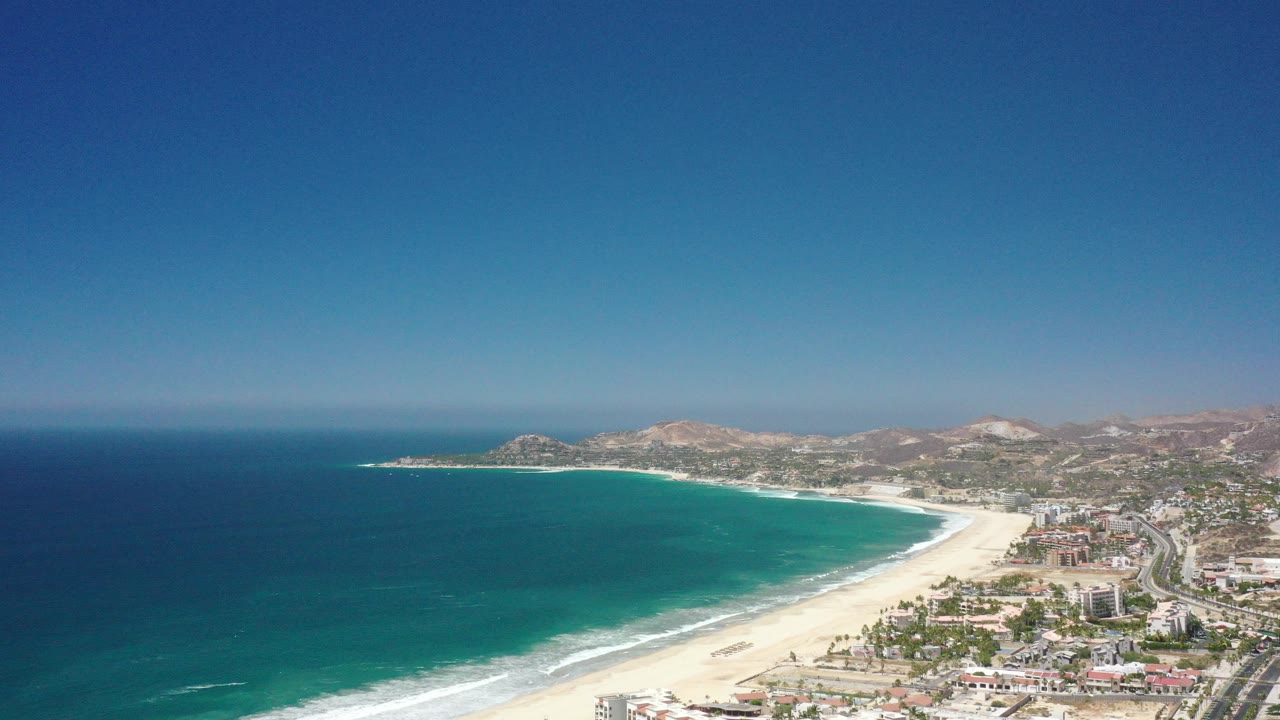 Wide aerial frame over San Jose Del Cabo, Mexico, with deep blue ocean at left, a pale curved beach, coastal buildings, dry hills, clear sky, and no visible people.