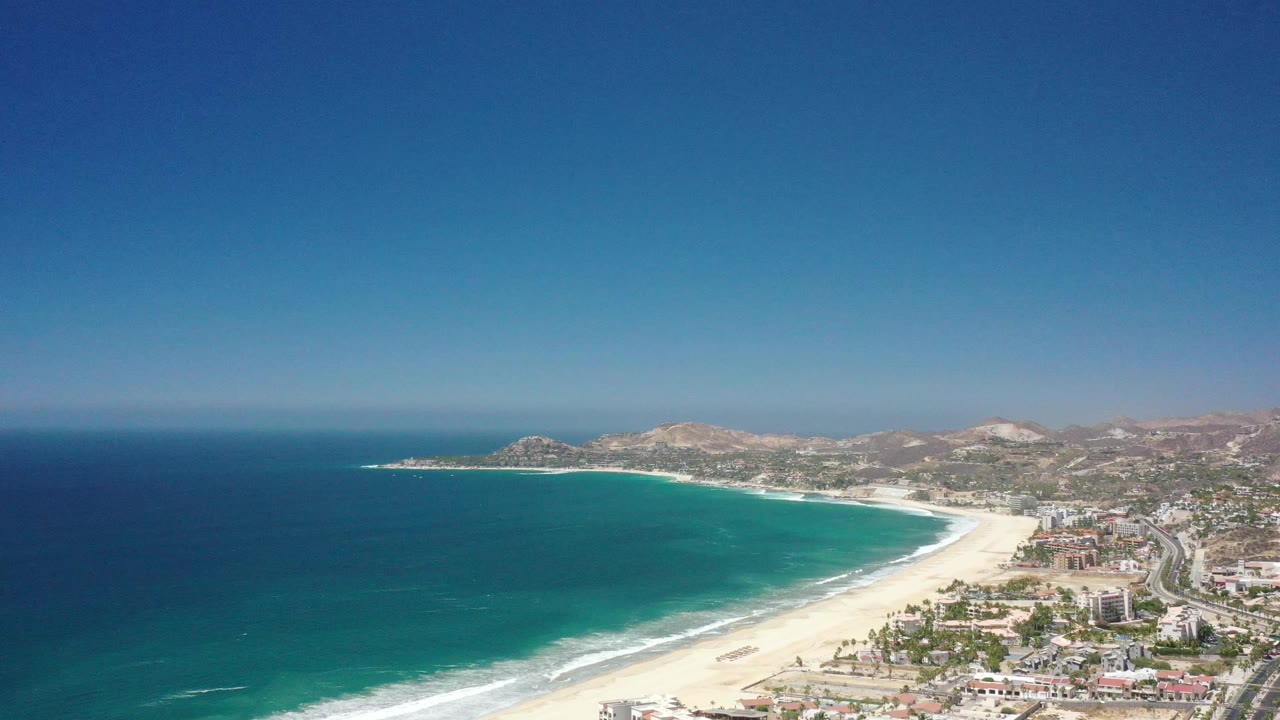 Coastal aerial with broad ocean, white beach, city edge, and hills.