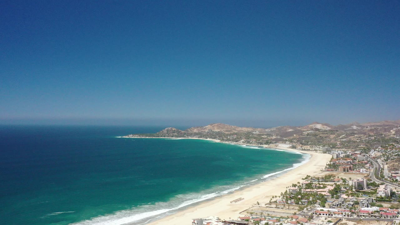 Wide aerial view over San Jose del Cabo, Mexico, with a curved sandy beach, blue ocean to the left, coastal buildings to the right, distant hills, and no visible people.