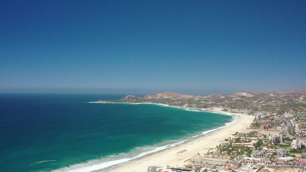 Aerial coastline with broad beach, ocean gradient, and hillside town.