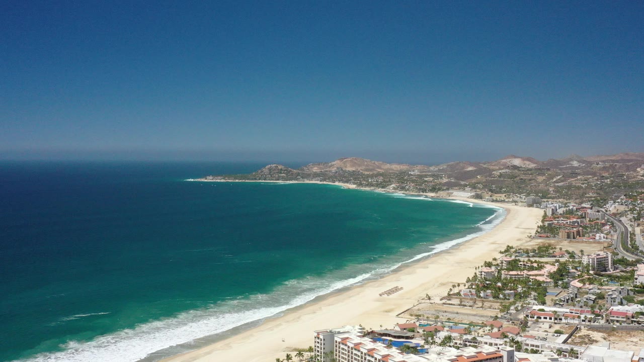 Aerial view of curved beach, turquoise sea, coastal buildings, and dry hills.
