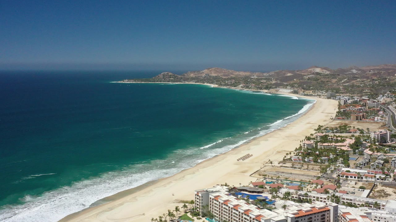 Aerial view of blue green ocean, sandy beach, coastal buildings, and dry hills.