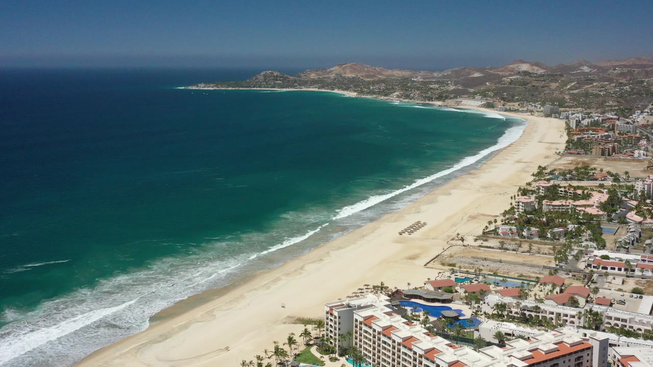 Aerial frame over San Jose Del Cabo showing teal ocean at left, a wide sandy beach curving through center, white surf, resort pools and buildings at lower right, and distant hills beyond the coast.