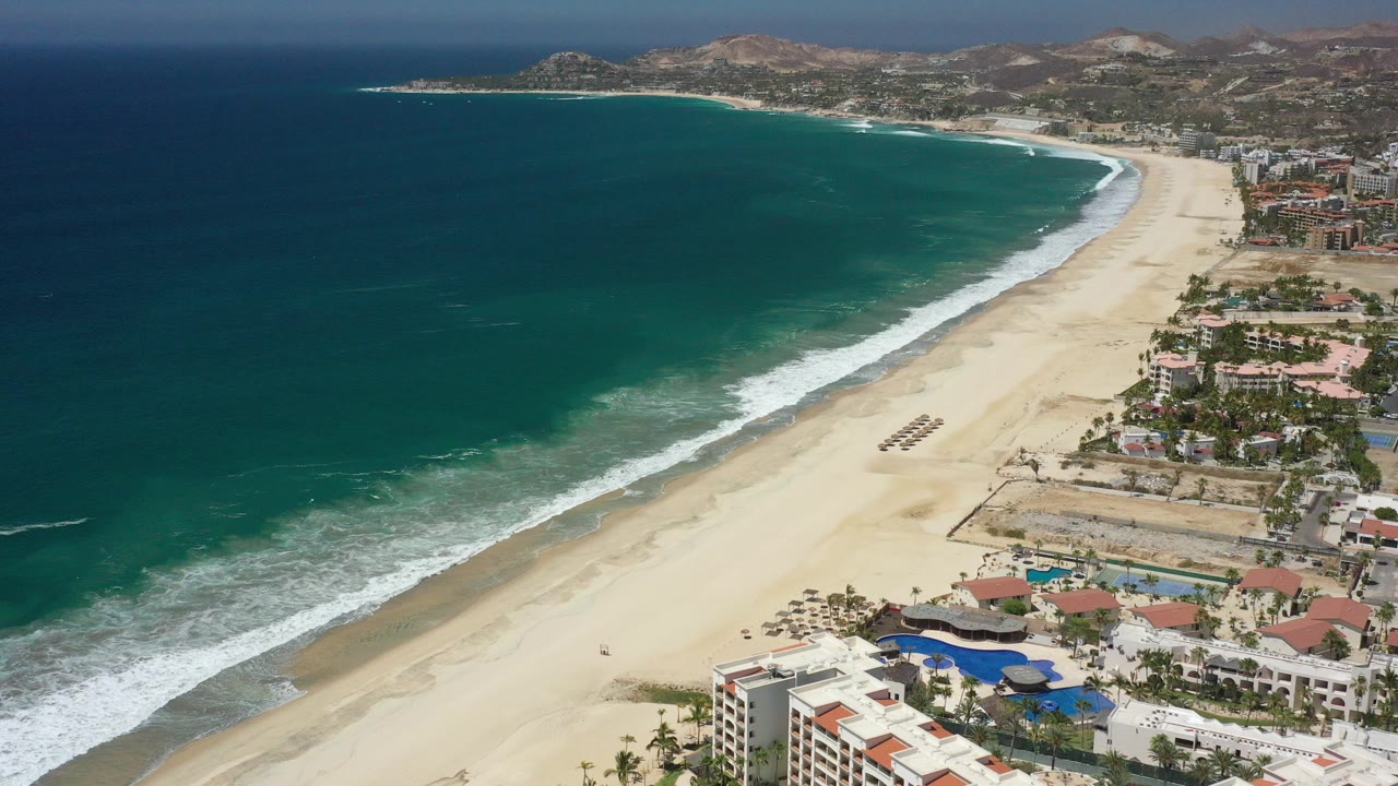 Aerial coastline with blue-green ocean, sandy beach, resorts, and dry hills.