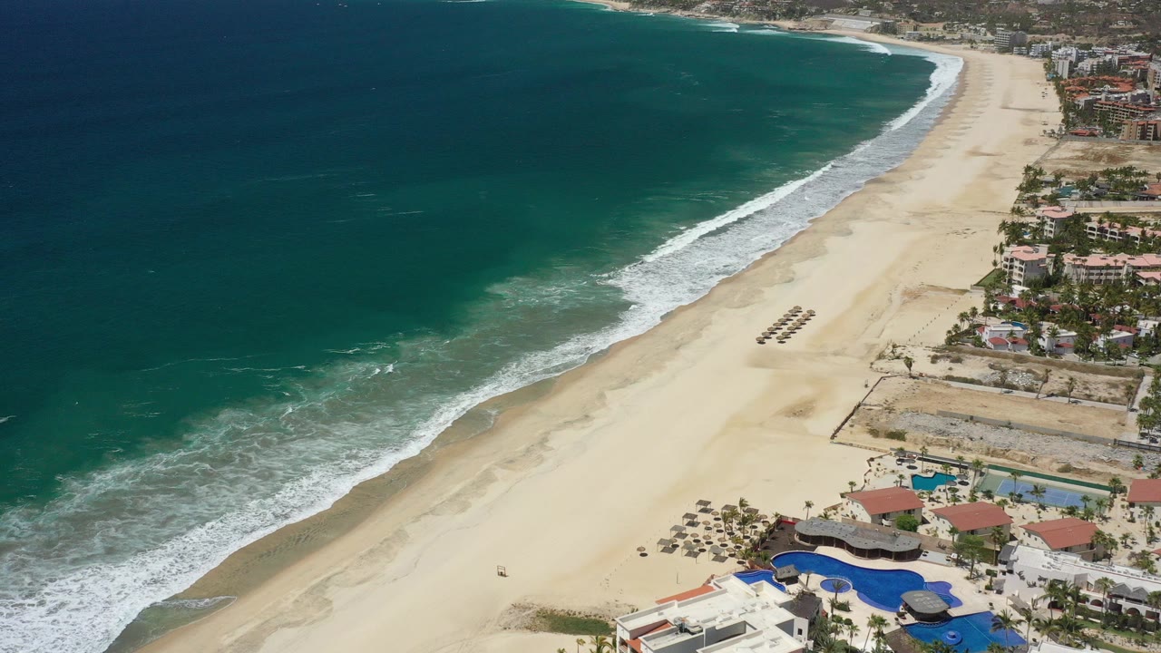 Opening aerial frame over San Jose Del Cabo, Mexico, showing teal water, a pale sandy beach, surf lines, pools, and coastal buildings with no visible people.