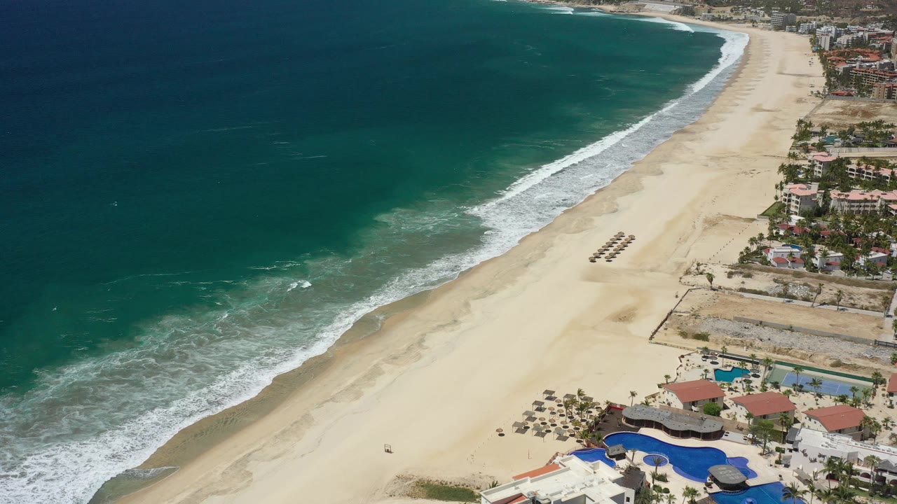 San Jose Del Cabo aerial beach frame showing white surf bands, open tan sand, a row of shade structures, pools, and clustered coastal buildings with no visible people.