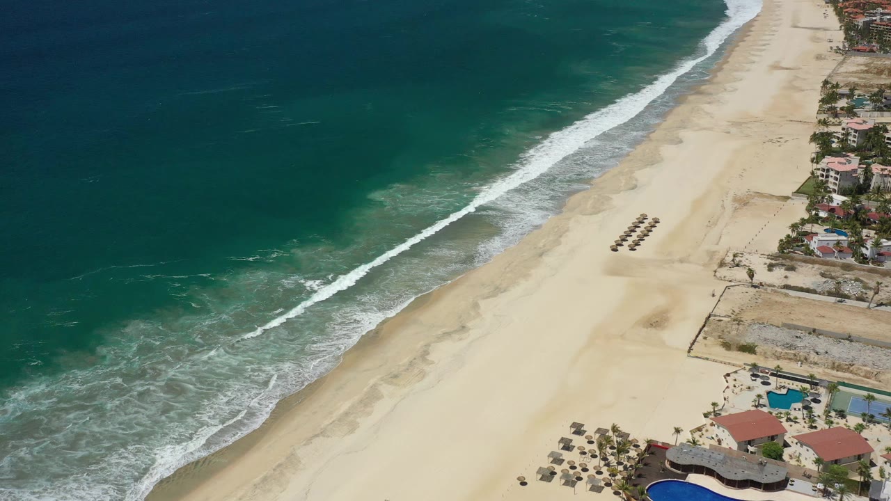 San Jose Del Cabo beach seen from above, with a long white surf band, open sand, clustered loungers near mid-beach, buildings at the far right, and no visible people.
