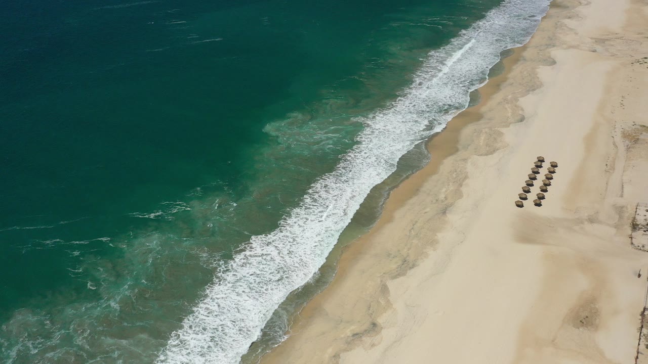 Aerial frame over San Jose del Cabo shoreline with teal water on the left, a white surf band across the beach, pale sand on the right, clustered umbrellas, and no visible people.