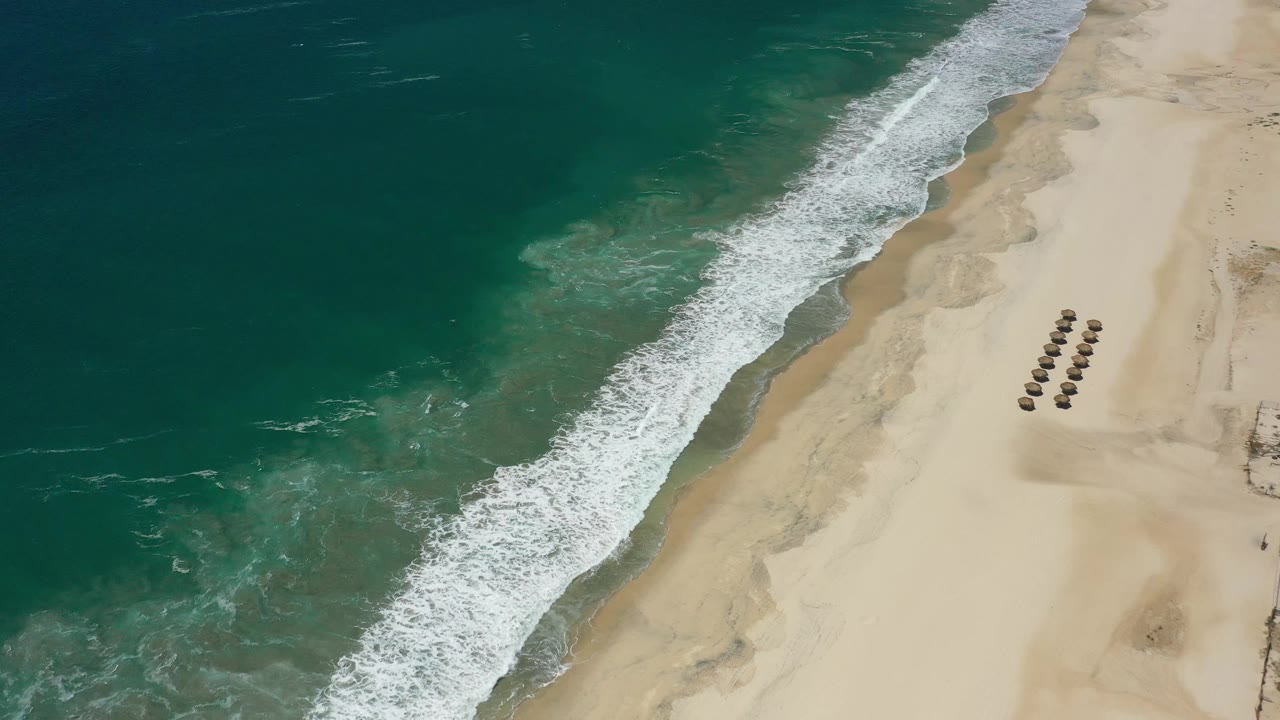 Overhead San Jose del Cabo beach view with the surf line shifted along the sand, darker teal water filling the left half, umbrella rows near the right edge, and no visible people.
