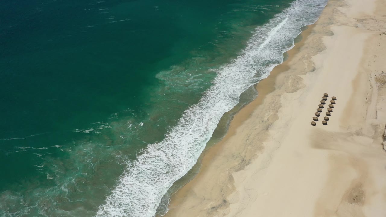Drone view of ocean surf beside open sand and a small umbrella cluster.