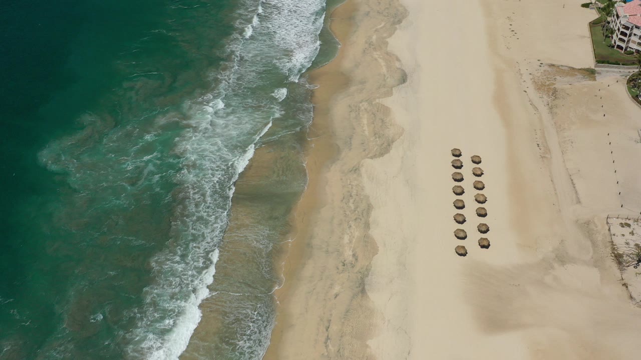 A high aerial frame over San Jose Del Cabo shows teal surf on the left, a broad sand beach, two neat rows of umbrellas, partial beachfront buildings, and tiny people visible on the sand.