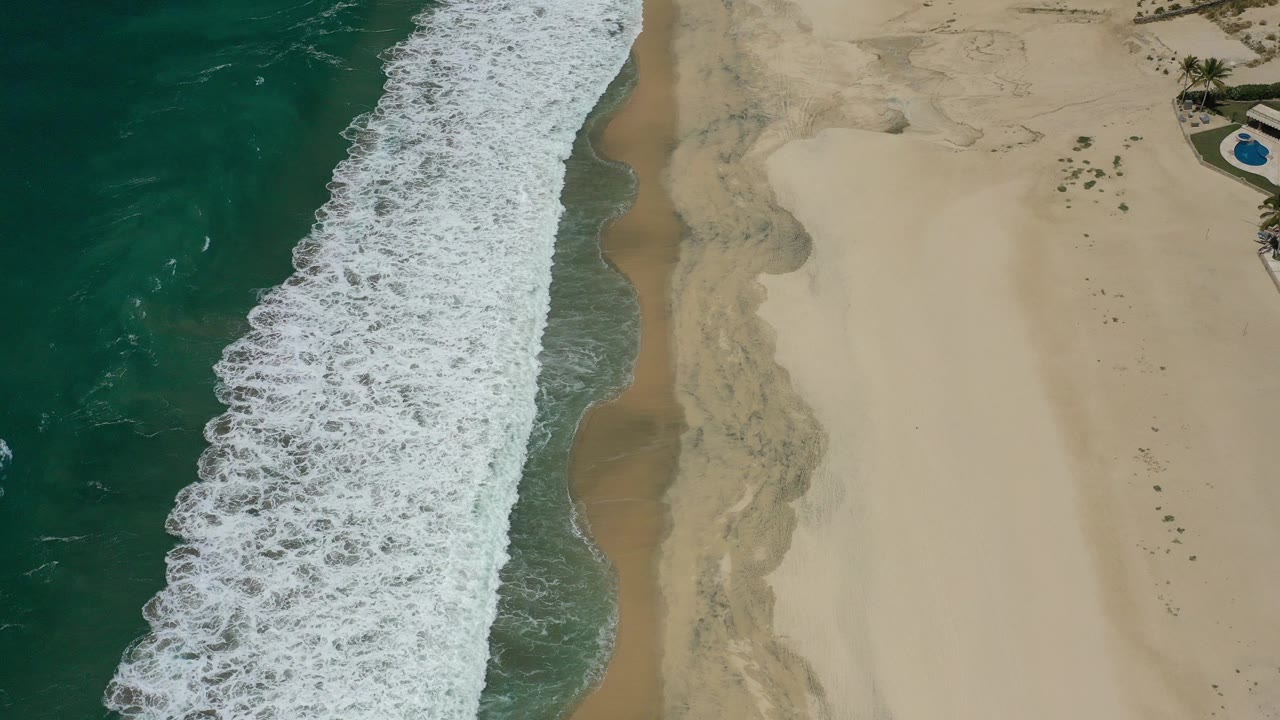 Overhead coastal frame from San Jose Del Cabo, Mexico, with dark teal water on the left, a white surf band, pale beach on the right, and no visible people.