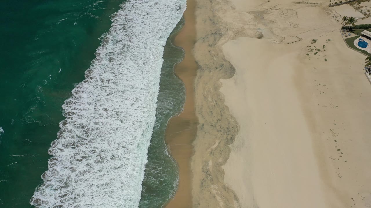 San Jose Del Cabo aerial view with a thick white foam line pushing up the beach, darker wet sand beside it, open pale sand, and no visible people.