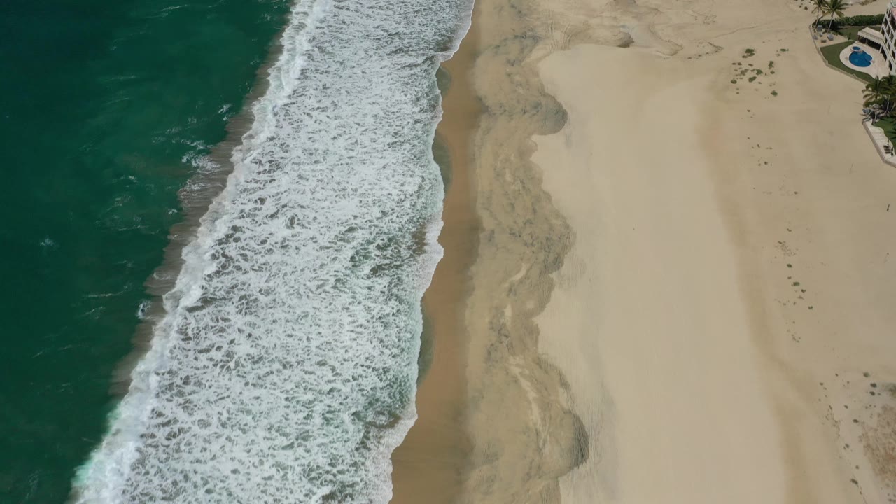 The San Jose Del Cabo shoreline fills the frame with foamy waves running diagonally, dry sand occupying the right half, faint wet sand patterns near the waterline, and no visible people.