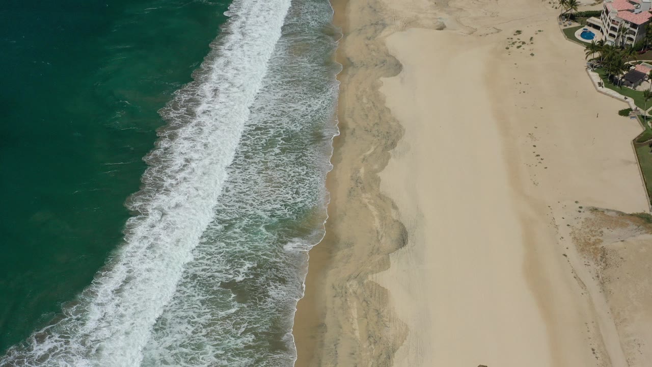 Aerial view of surf meeting a sandy beach beside resort pools.