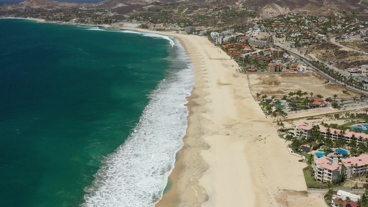 Aerial view of teal ocean, white surf, sandy beach, coastal buildings, and dry hills.