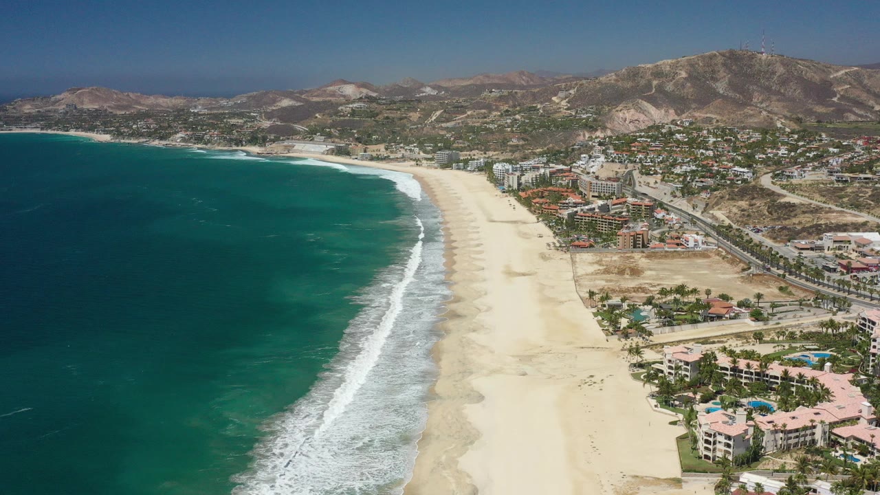 High aerial view over San Jose del Cabo, Mexico, with turquoise water on the left, a white surf line, a broad sand beach, coastal buildings, roads, and dry hills in the background, no visible people.