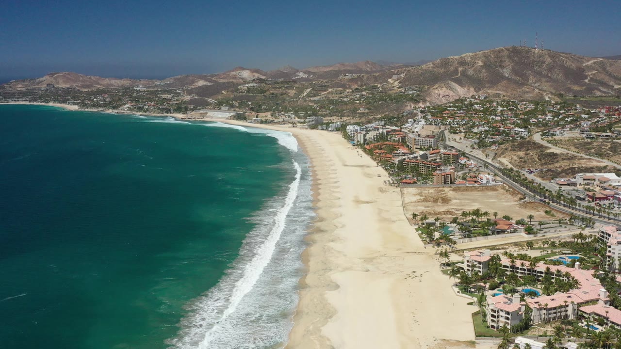 Drone-like view of surf, open sand, nearby buildings, roads, and mountains.