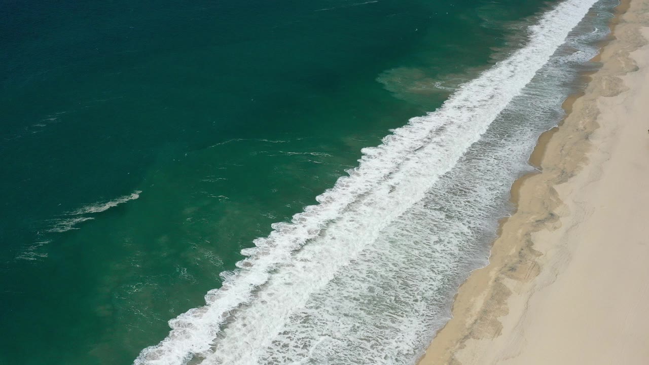 Aerial view of green ocean waves breaking onto pale sand.