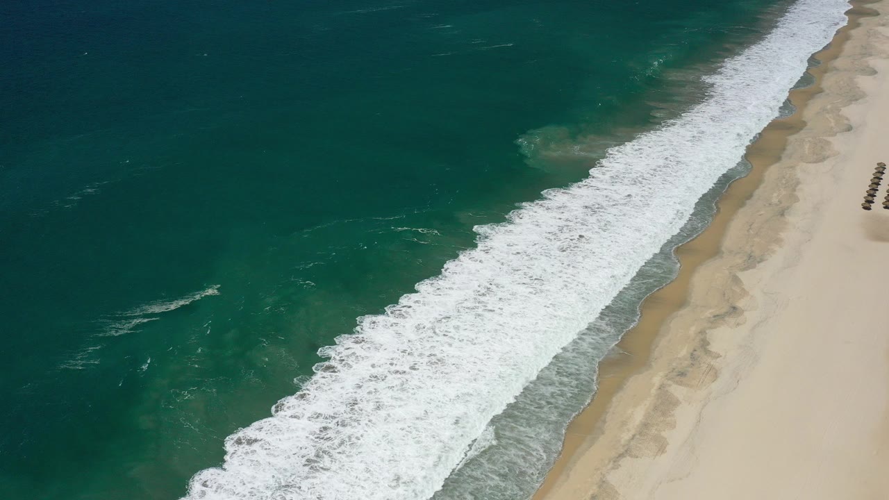 High oblique aerial view of blue-green water and white surf running diagonally along a San Jose del Cabo beach, with pale sand at right and no visible people.