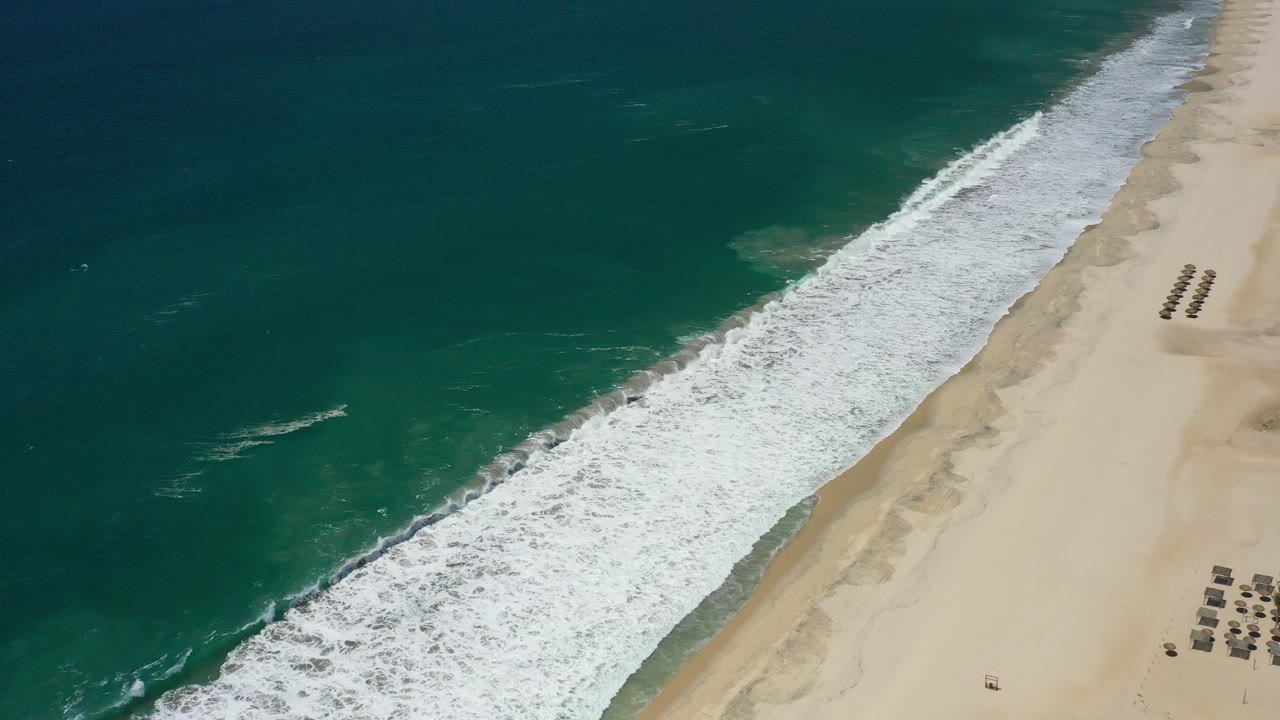 Aerial frame over the San Jose del Cabo coast with deep blue-green water, a diagonal band of white surf, pale sand, beach furniture at the far right, and no visible people.