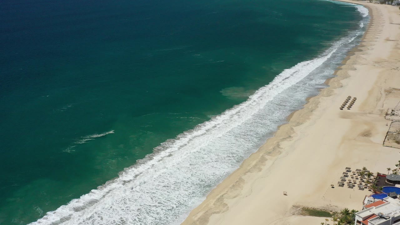 High aerial view over San Jose del Cabo with teal water on the left, white surf crossing a pale sand beach, resort buildings at the lower right edge, and no visible people.