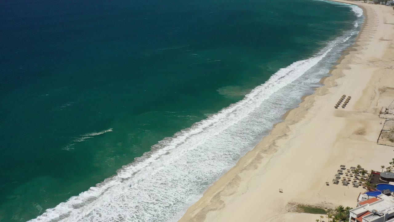 Aerial frame over San Jose del Cabo showing a diagonal band of breaking surf, dark teal open water, pale beach, organized beach umbrellas or loungers, and no visible people.