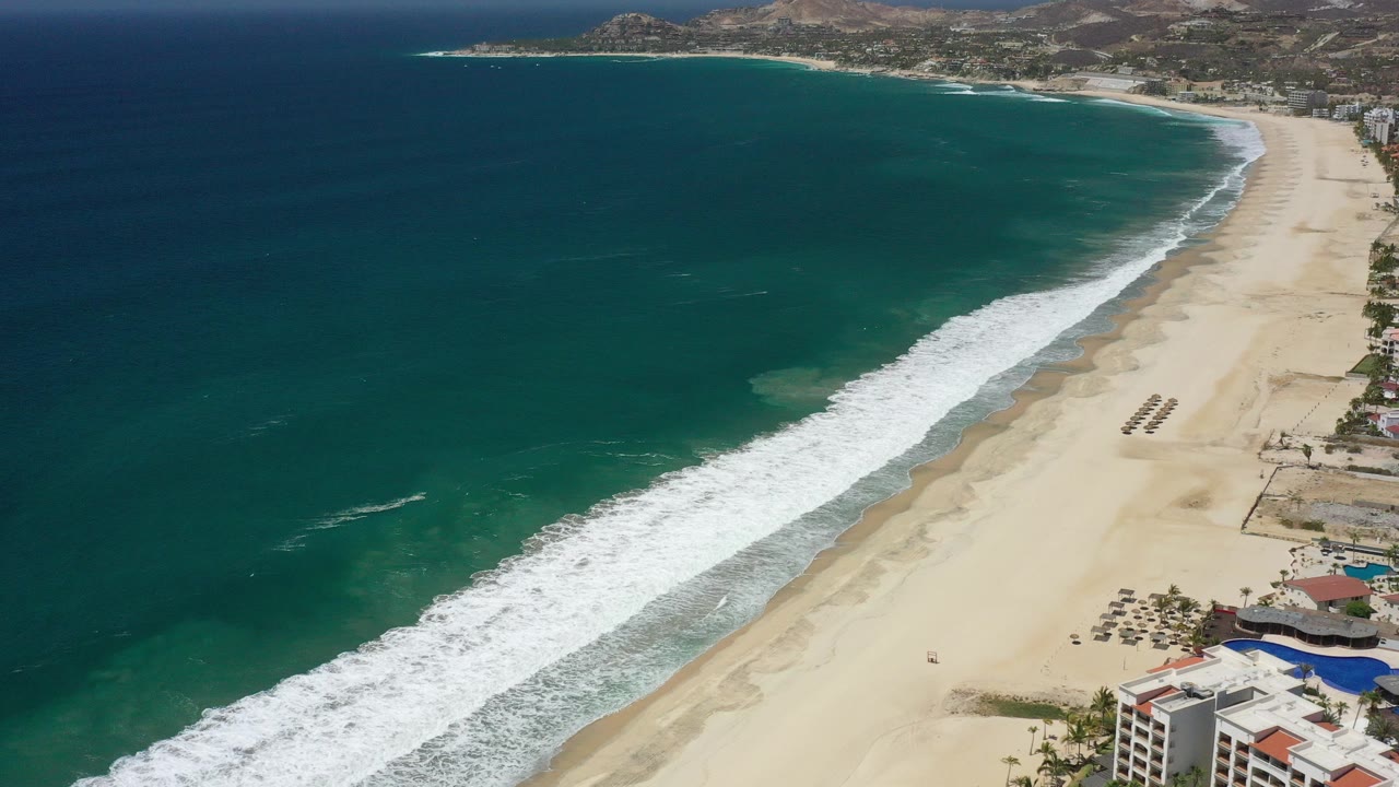 Aerial view over San Jose del Cabo, Mexico, with turquoise ocean filling the left side, white surf reaching a long sandy beach, coastal buildings at lower right, and no visible people.