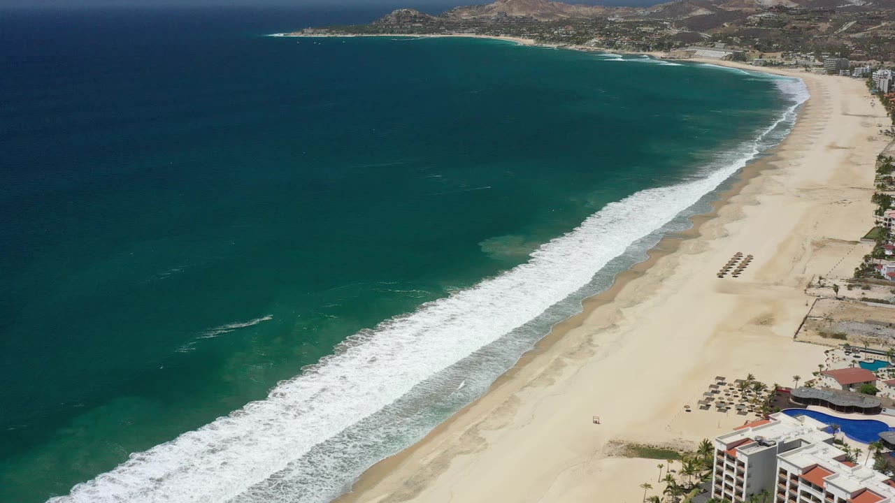 Aerial frame over San Jose del Cabo, Mexico, showing a diagonal surf band, deep blue-green water, a pale beach curving toward distant hills, and no visible people.