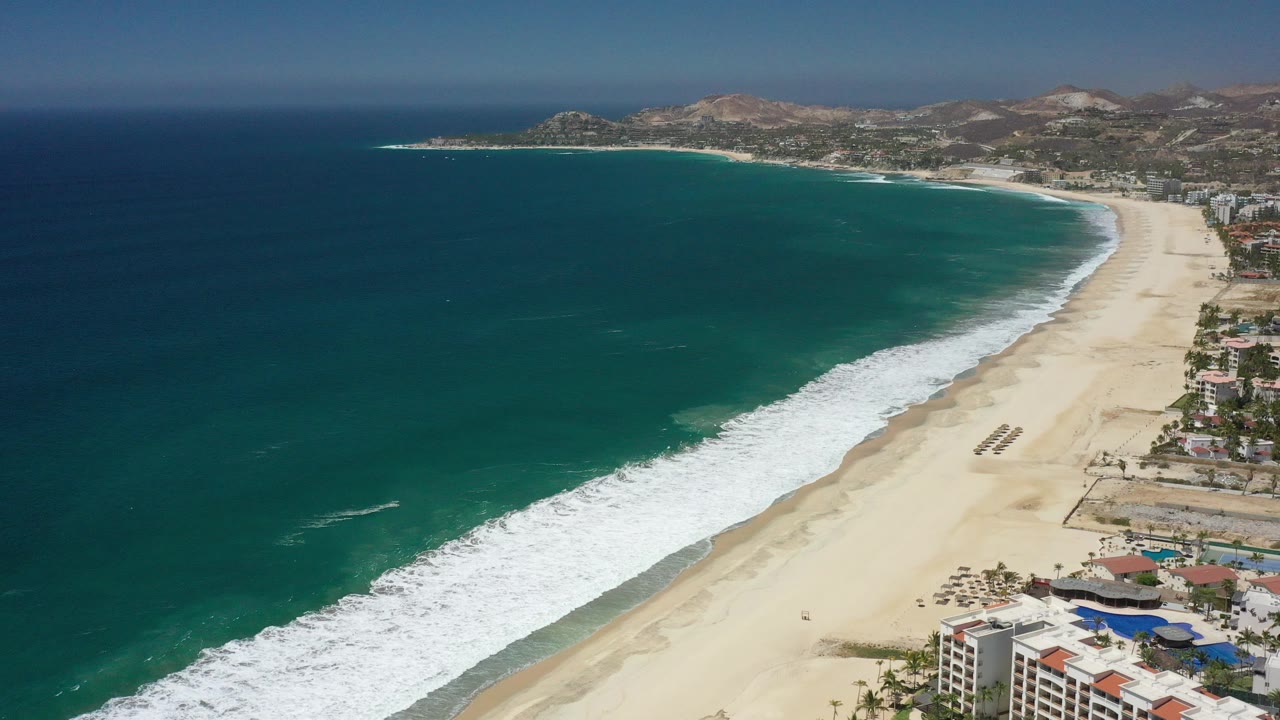 Aerial coastline with teal ocean, white surf, sandy beach, buildings, and hills.