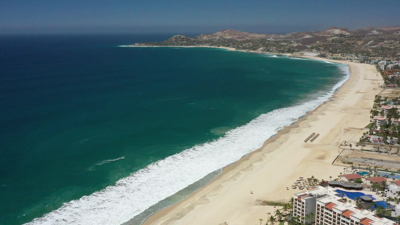 Aerial view one second into the San Jose Del Cabo clip, with the surf band slightly farther along the beach, coastal buildings at lower right, hills beyond, and no visible people.