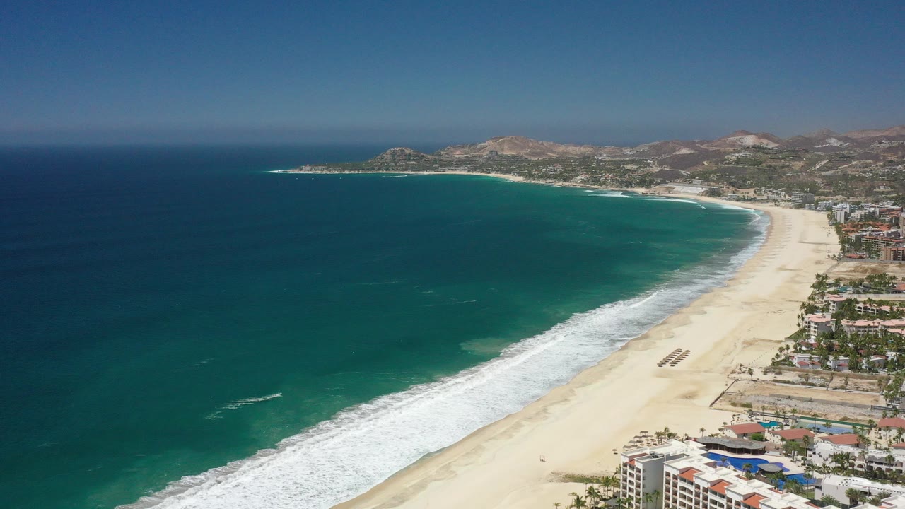 Aerial view of turquoise ocean beside a wide sandy beach and beachfront buildings.