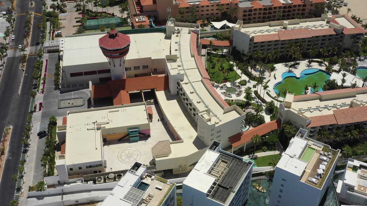 High aerial frame over San Jose Del Cabo resort buildings, centered on a red saucer-shaped tower beside cream rooftops, palm landscaping, turquoise pools, a road, and no visible people.