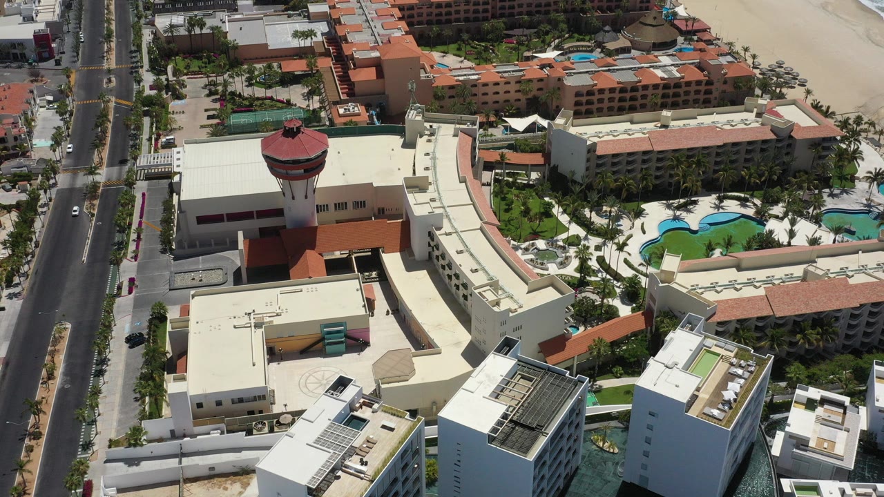 High aerial frame over San Jose del Cabo resort buildings, a round red-roof tower, pools, a coastal road, sandy beach, and surf, with no visible people.