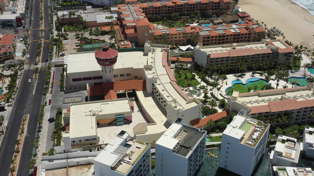 Aerial coastal resort view with tower, hotel roofs, pools, road, and shoreline.