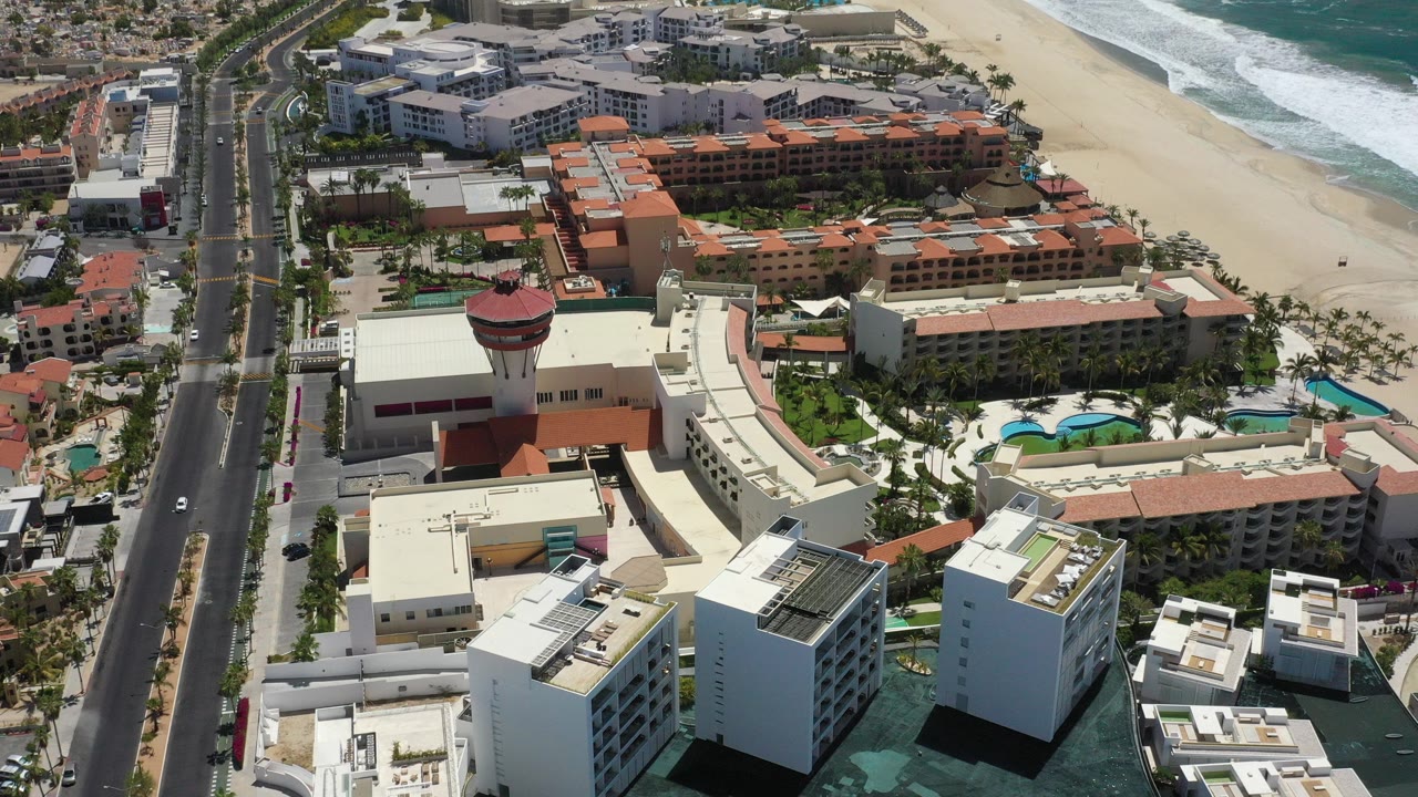 Aerial frame over San Jose del Cabo showing a divided road, resort buildings with red tile roofs, pools, pale beach, turquoise ocean, white surf, and no visible people.