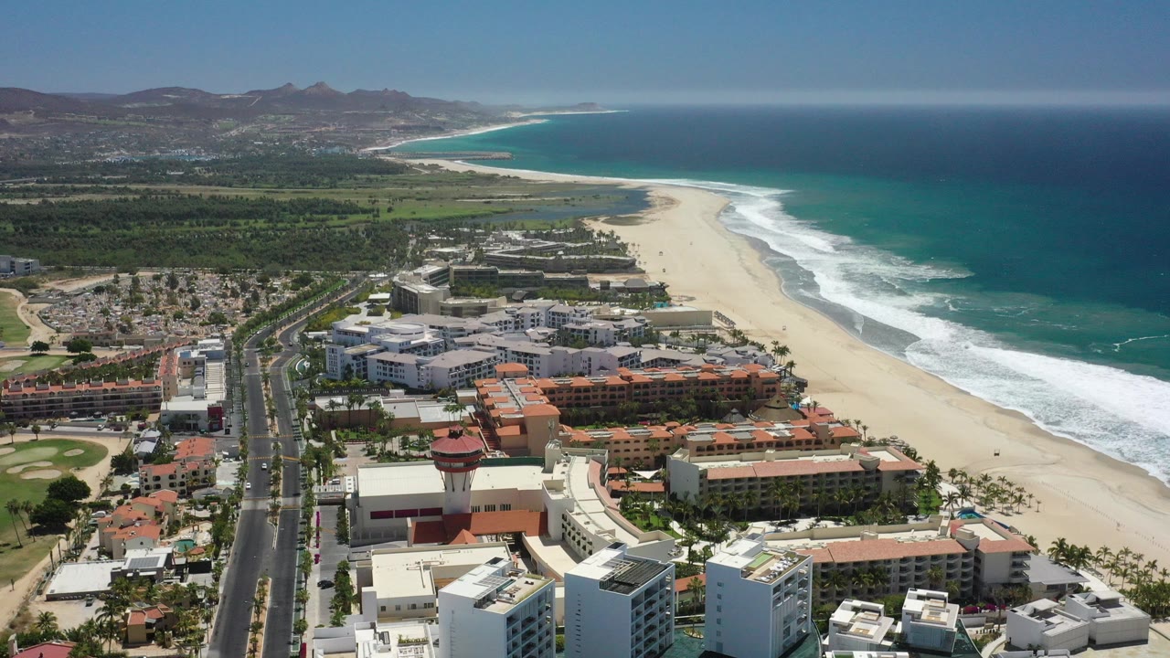 High aerial view of beachfront buildings, road, sandy beach, surf, ocean, and mountains.