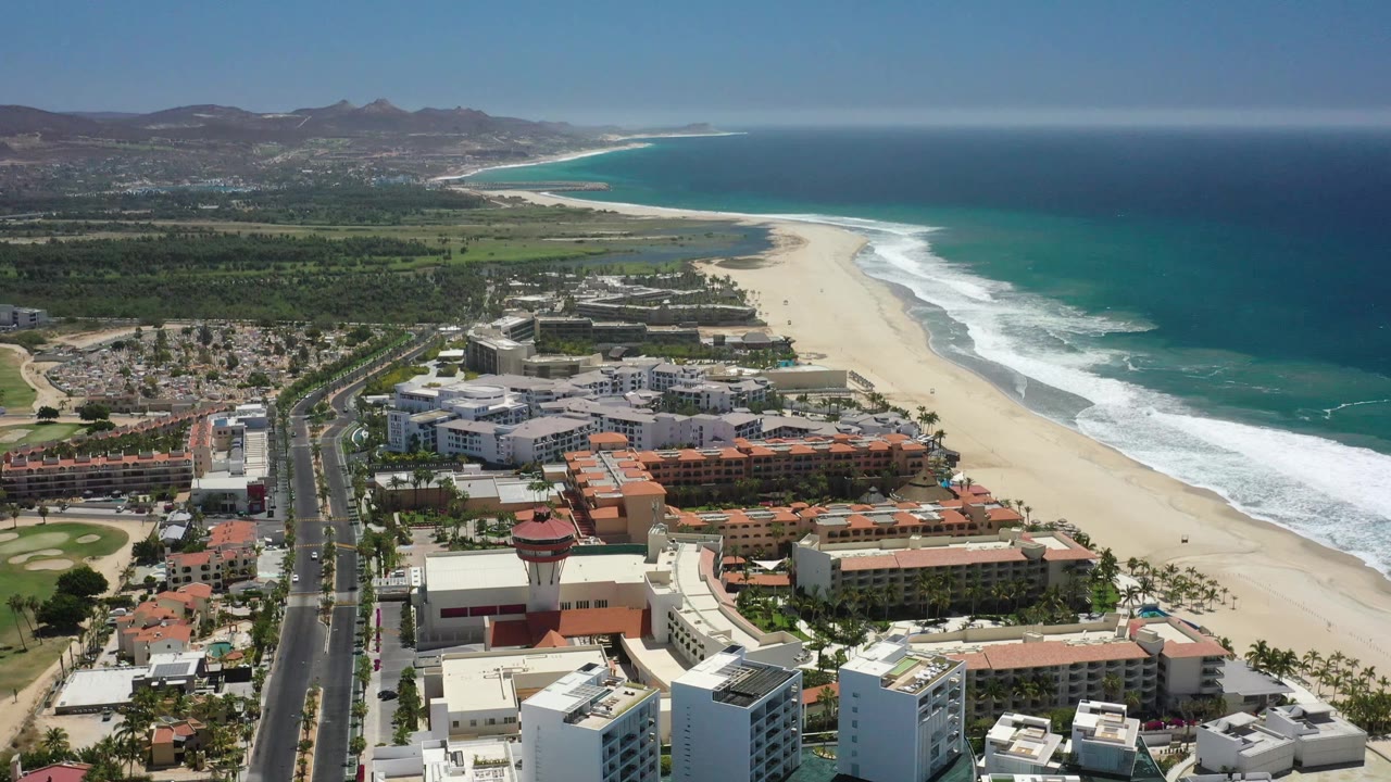 Drone perspective over San Jose del Cabo showing the road and hotel blocks slightly farther along the coast, with breaking waves edging the beach and no visible people.