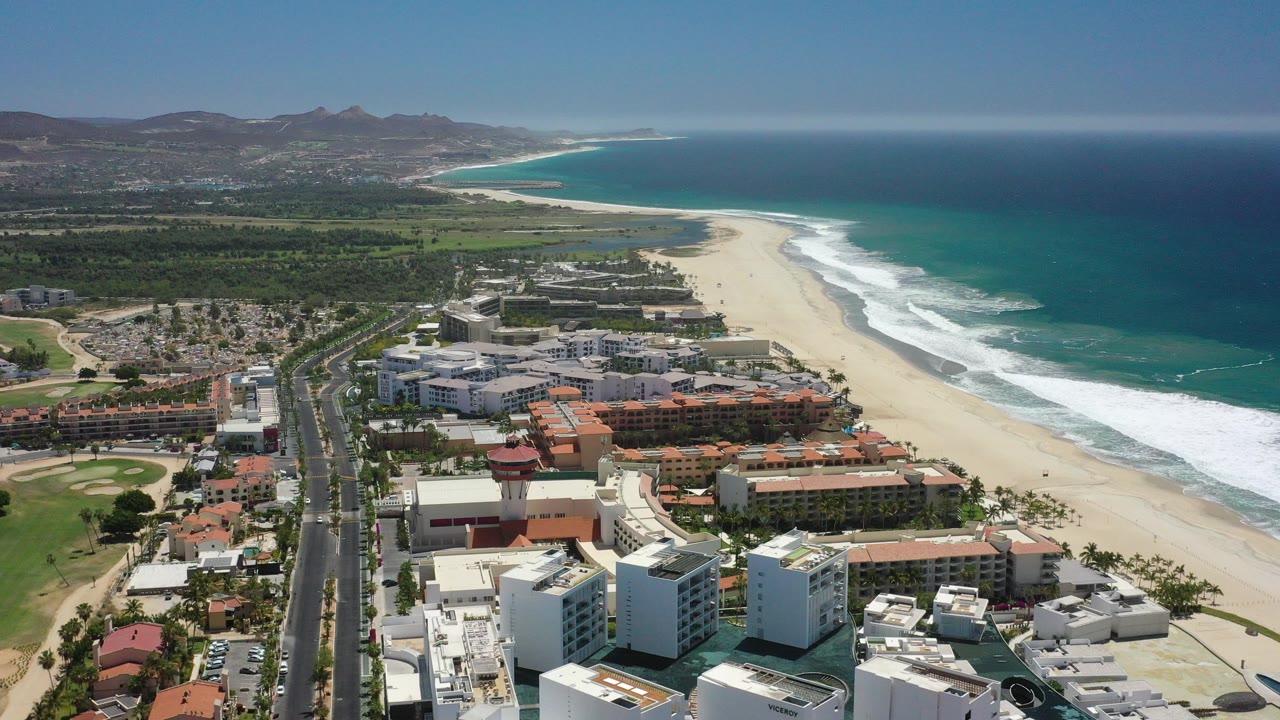 Aerial view of beachfront buildings, road, beach, surf, ocean, and mountains.