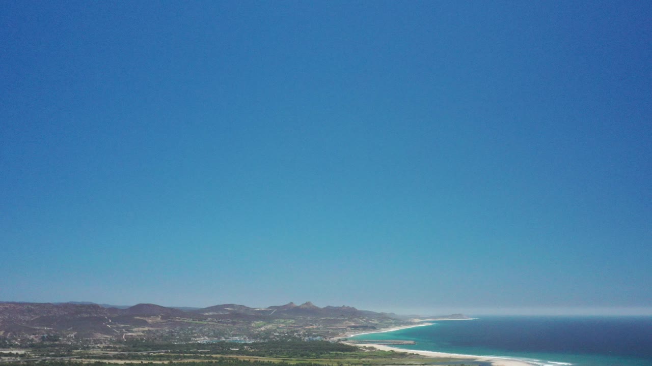 Aerial coastline with blue ocean, sandy beach, town, green land, hills, and clear sky.
