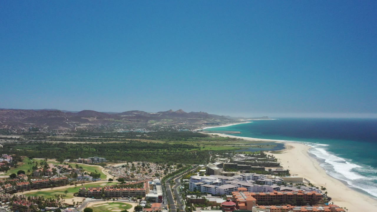 High aerial view over San Jose del Cabo with resort buildings near the lower edge, a pale beach and surf line to the right, blue ocean beyond, inland greenery and brown hills in the distance, and no visible people.
