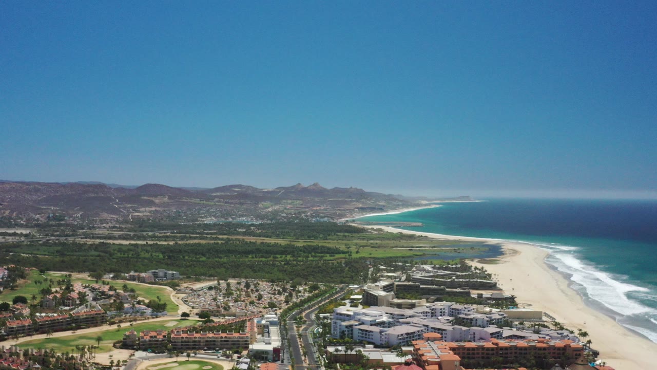 High aerial view over San Jose del Cabo with beachfront buildings and roads below, a wide pale beach, turquoise ocean, white surf, inland greenery, distant brown hills, and no visible people.