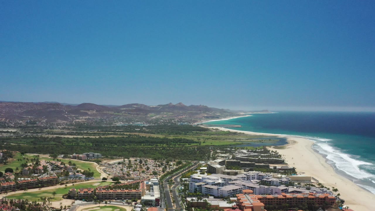Wide aerial coastal view with beach hotels, road crossing area, surf, ocean, and hills.
